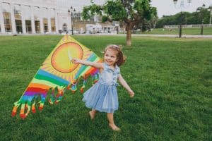 Joyful little cute child baby girl in denim dress walking, play with colorful kite and have fun in green park