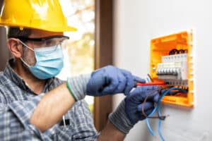 Electrician at work on an electrical panel protected by helmet, safety goggles and gloves; wear the surgical mask to prevent the spread of Coronavirus. Construction industry. Covid-19 Prevention.